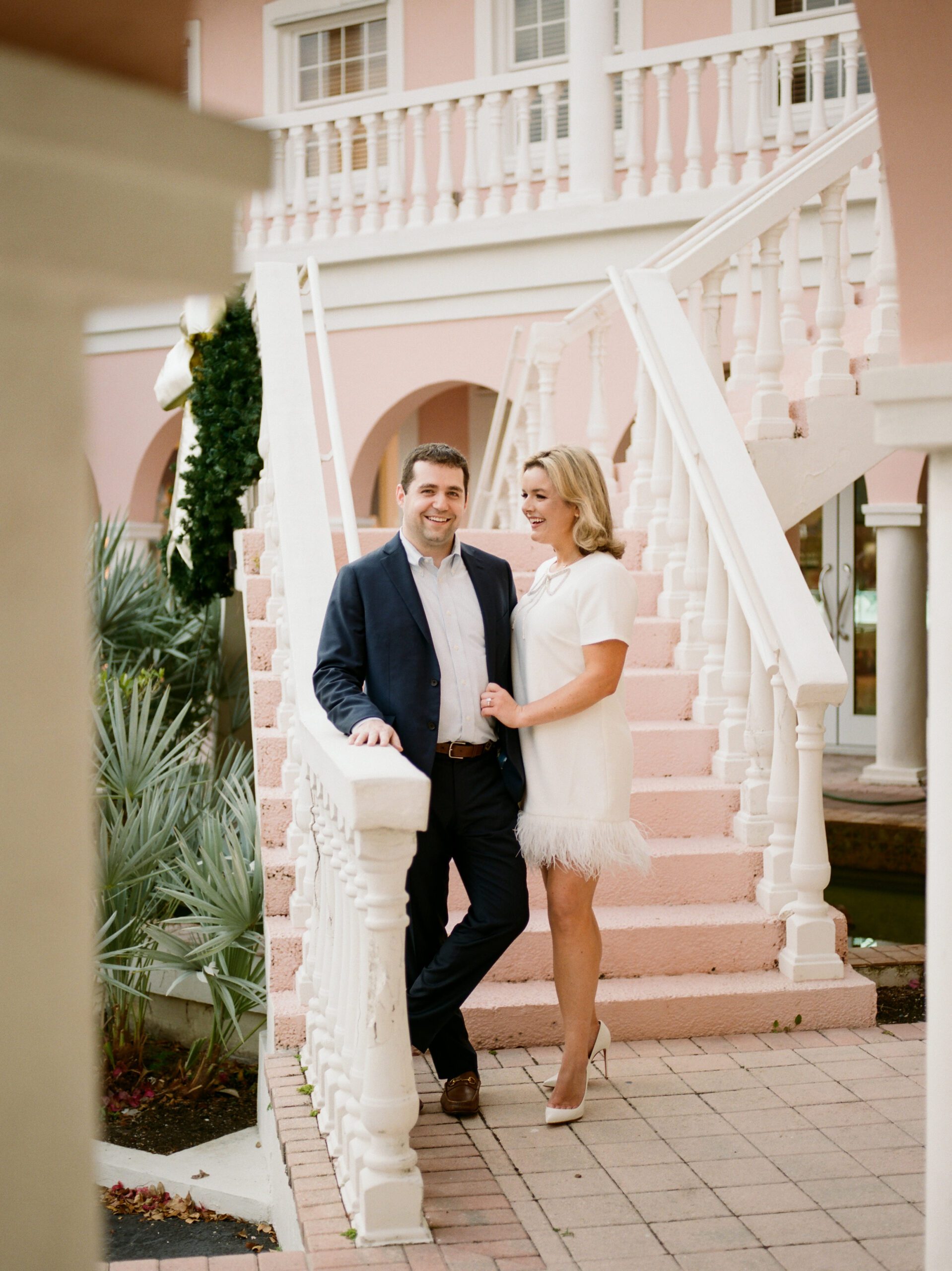 An engagement photo taken in downtown old naples with a pink staircase building and a couple standing in front of it looking lovingly at each other